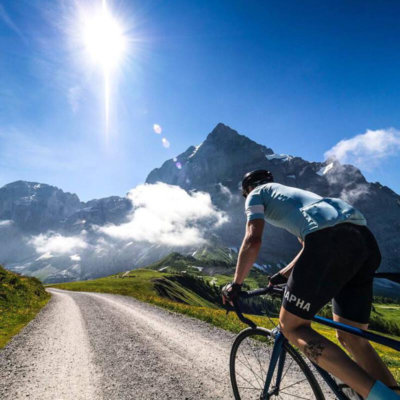 This may contain: a man riding a bike down a dirt road next to a lush green hillside covered in clouds